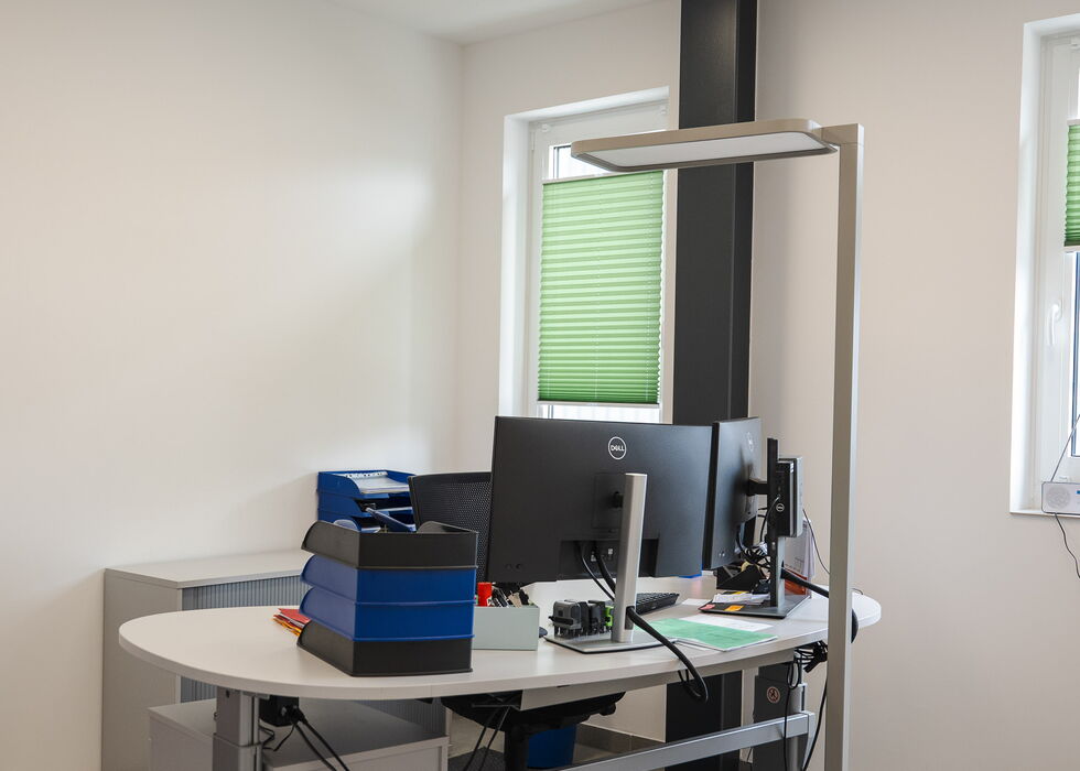 Ein moderner Büroarbeitsplatz mit gebogenem Schreibtisch, zwei Monitoren, einer Stehlampe und Fenstern mit grünen Jalousien.