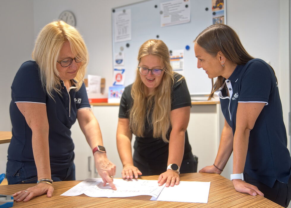 Three women in navy polos lean over papers on a table in an office or classroom, discussing the documents.