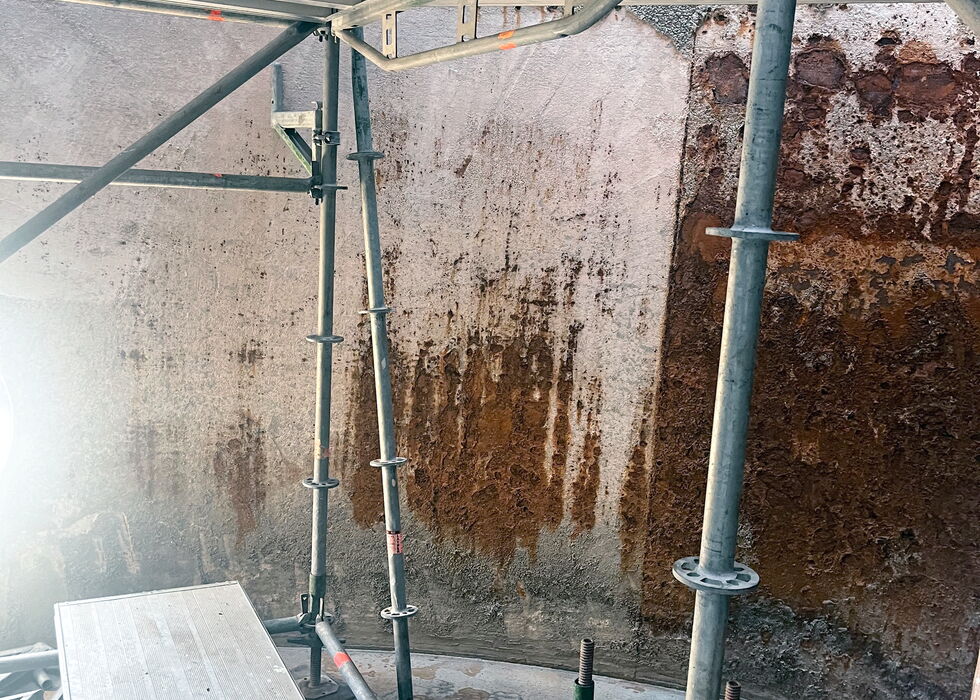 Metallgerüst und Arbeitsplattform in einem Behälter mit feuchten, fleckigen Wänden während der Sanierung.