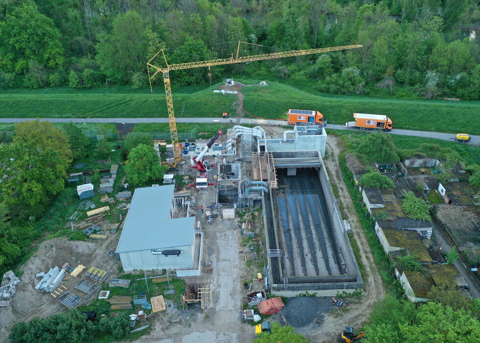 Luftaufnahme einer Baustelle mit Kran und Betonbauwerk neben einem Fluss in grüner Landschaft.
