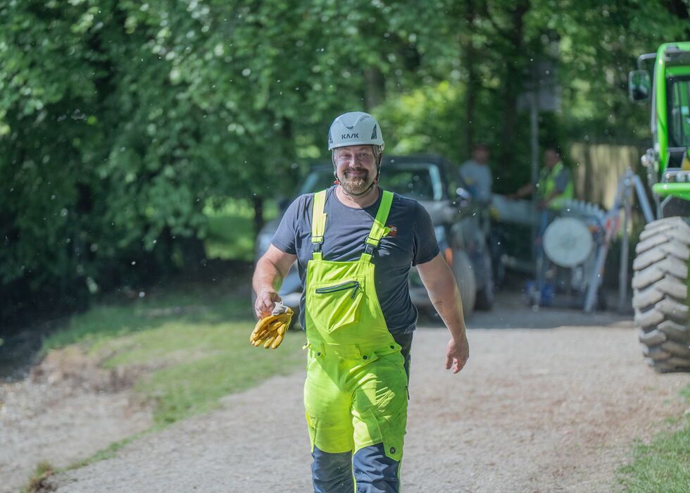 Ein Mann in neongrüner Latzhose und Helm geht auf einem Schotterweg neben Landmaschinen.