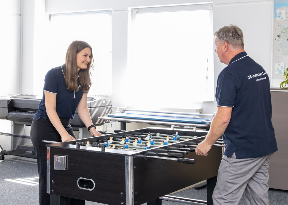 Zwei Kollegen bauen einen Kickertisch in einem hellen Büroraum auf.