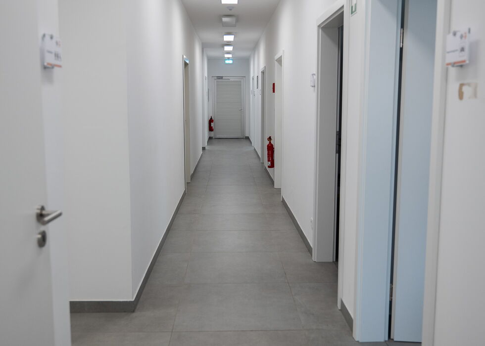 Langer, sauberer Flur mit weißen Wänden, grauem Fliesenboden, Deckenleuchten und offenen Türen rechts.