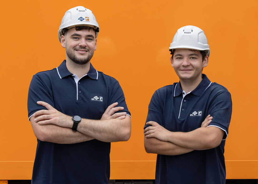 Zwei lächelnde Bauarbeiter mit weißen Schutzhelmen und navyblauen Poloshirts stehen mit verschränkten Armen vor orangefarbenem Hintergrund.