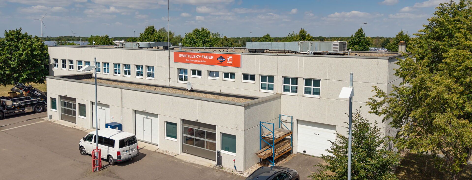 Weitwinkelansicht eines hellen Industriegebäudes mit einem Lieferwagen und einem Auto vor dem Gebäude unter blauem Himmel.