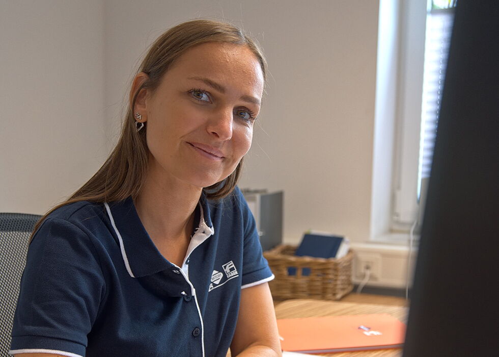 A woman in a navy polo shirt uses a mouse at her desk beside a monitor and laptop in an office.