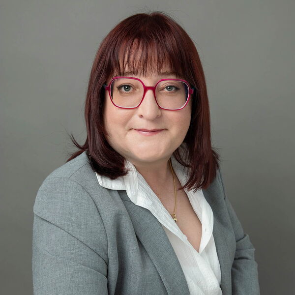 Porträt einer Frau mit pinker Brille und grauem Blazer vor neutralem Hintergrund.