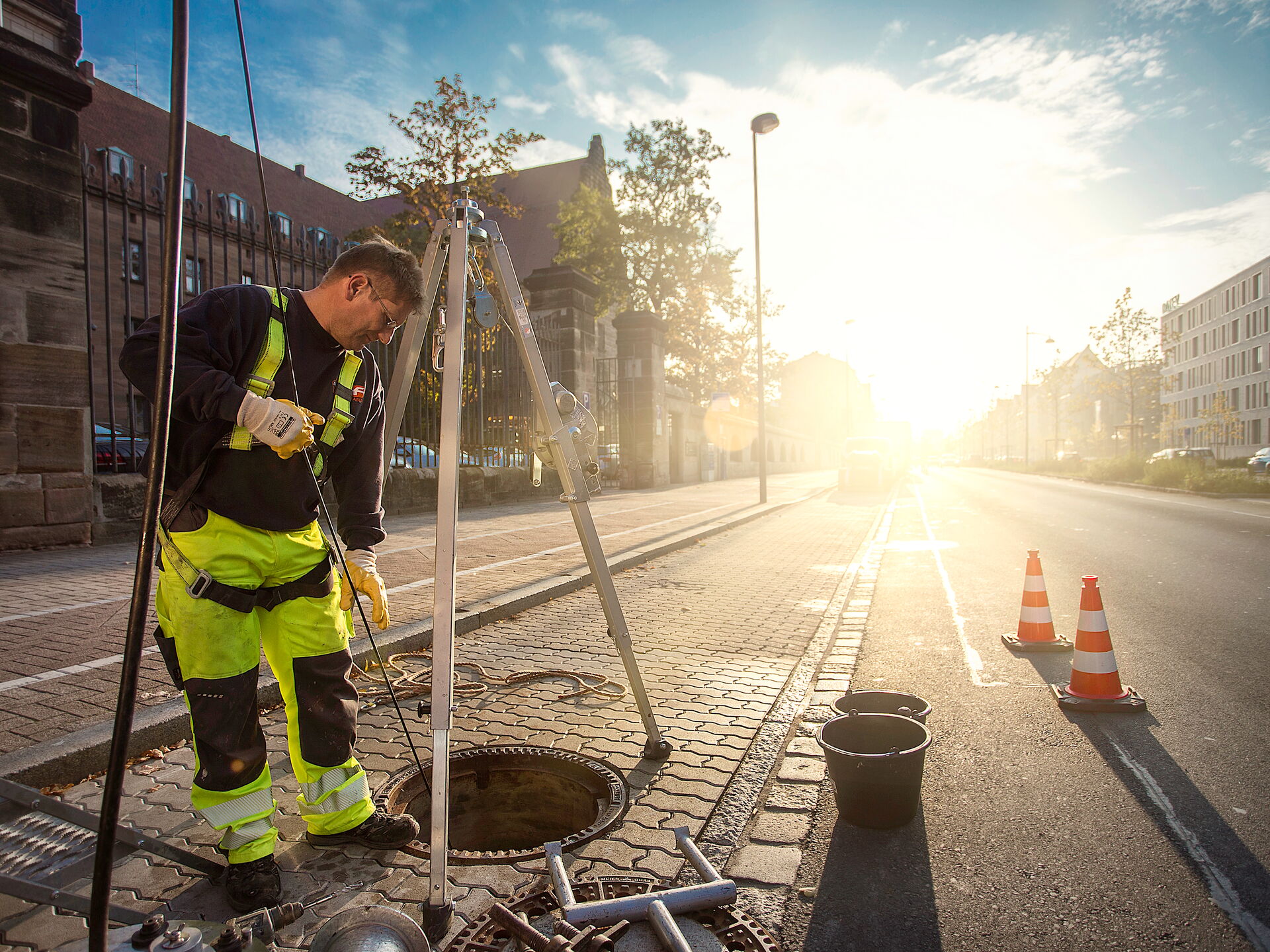 Ein Straßenarbeiter in Warnkleidung prüft einen offenen Schacht auf einer Stadtstraße bei Sonnenaufgang.