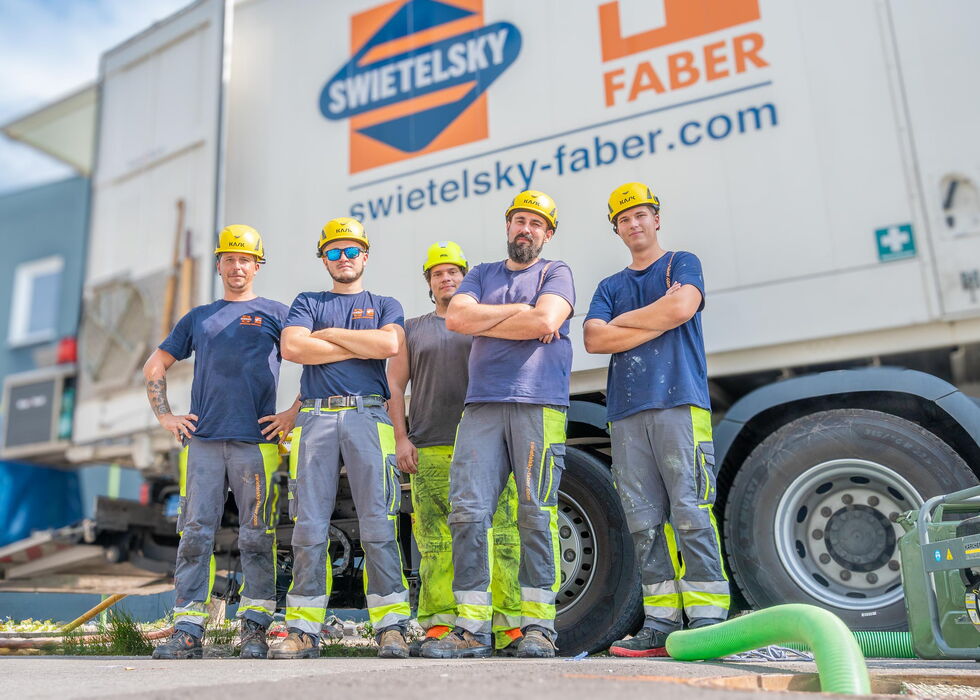 Fünf Bauarbeiter mit Helmen stehen auf einer Baustelle vor einem Fahrzeug von Swietelsky Faber.