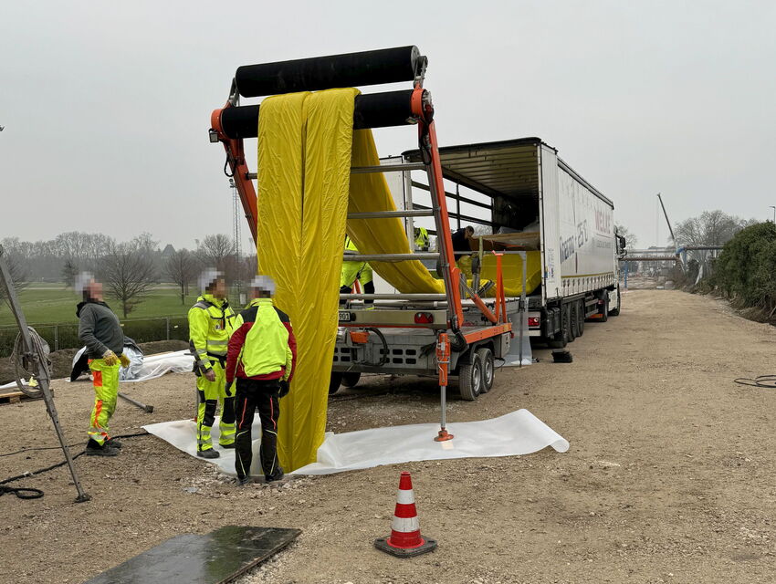 Arbeiter in Warnkleidung entladen eine große gelbe Geberolle neben einer Landstraße aus einem Lkw.