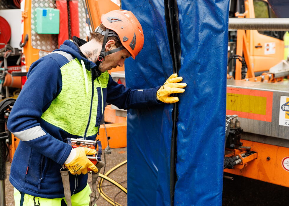 Ein Arbeiter mit Schutzkleidung richtet eine blaue Abdeckung neben orangefarbener Industrieanlage aus.