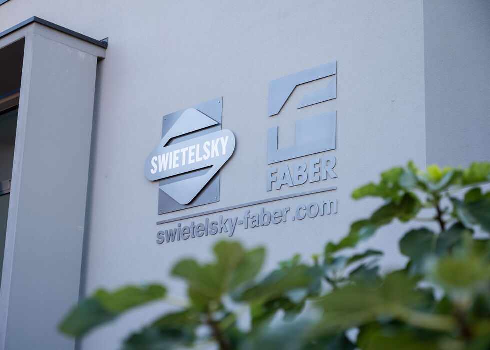 Nahaufnahme des SWIETELSKY-FABER Gebäudeschilds an der Fassade, im Vordergrund Grünpflanzen.
