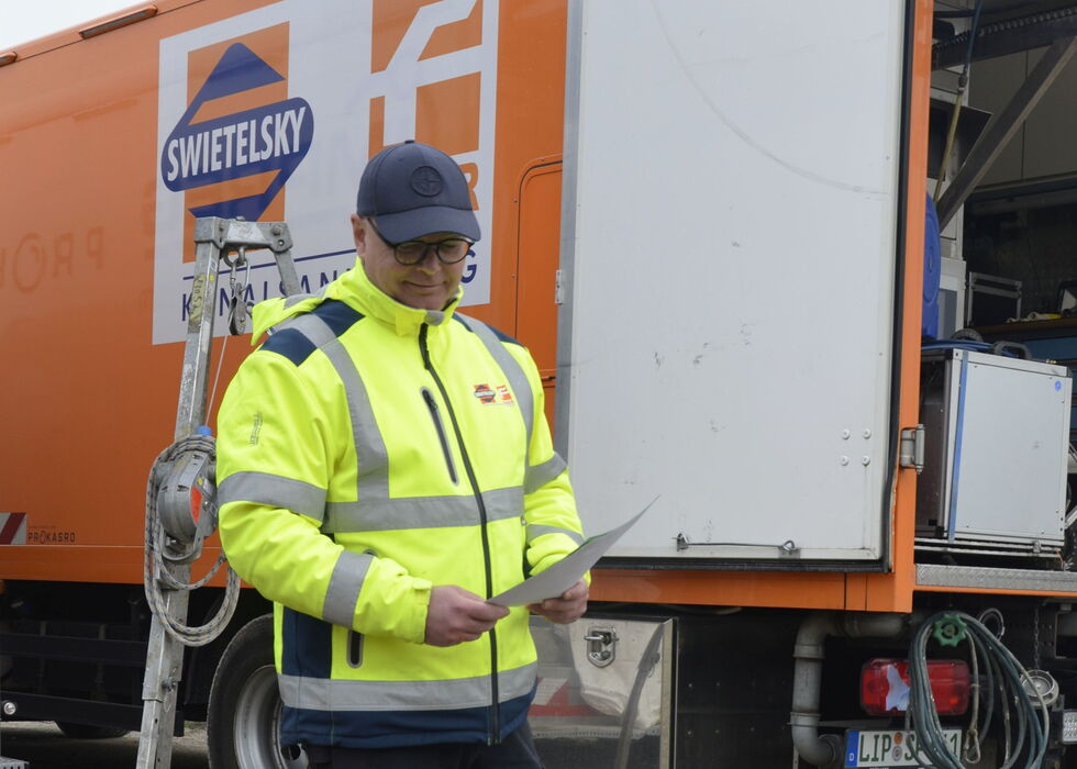 Ein Arbeiter in Warnjacke liest Notizen neben einem Servicewagen auf einer Baustelle.