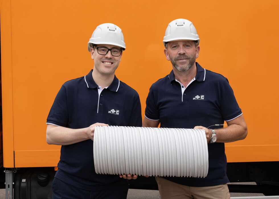 Zwei lächelnde Arbeiter mit Schutzhelmen halten einen großen weißen Schlauchaufroller vor einem orangefarbenen Lkw.