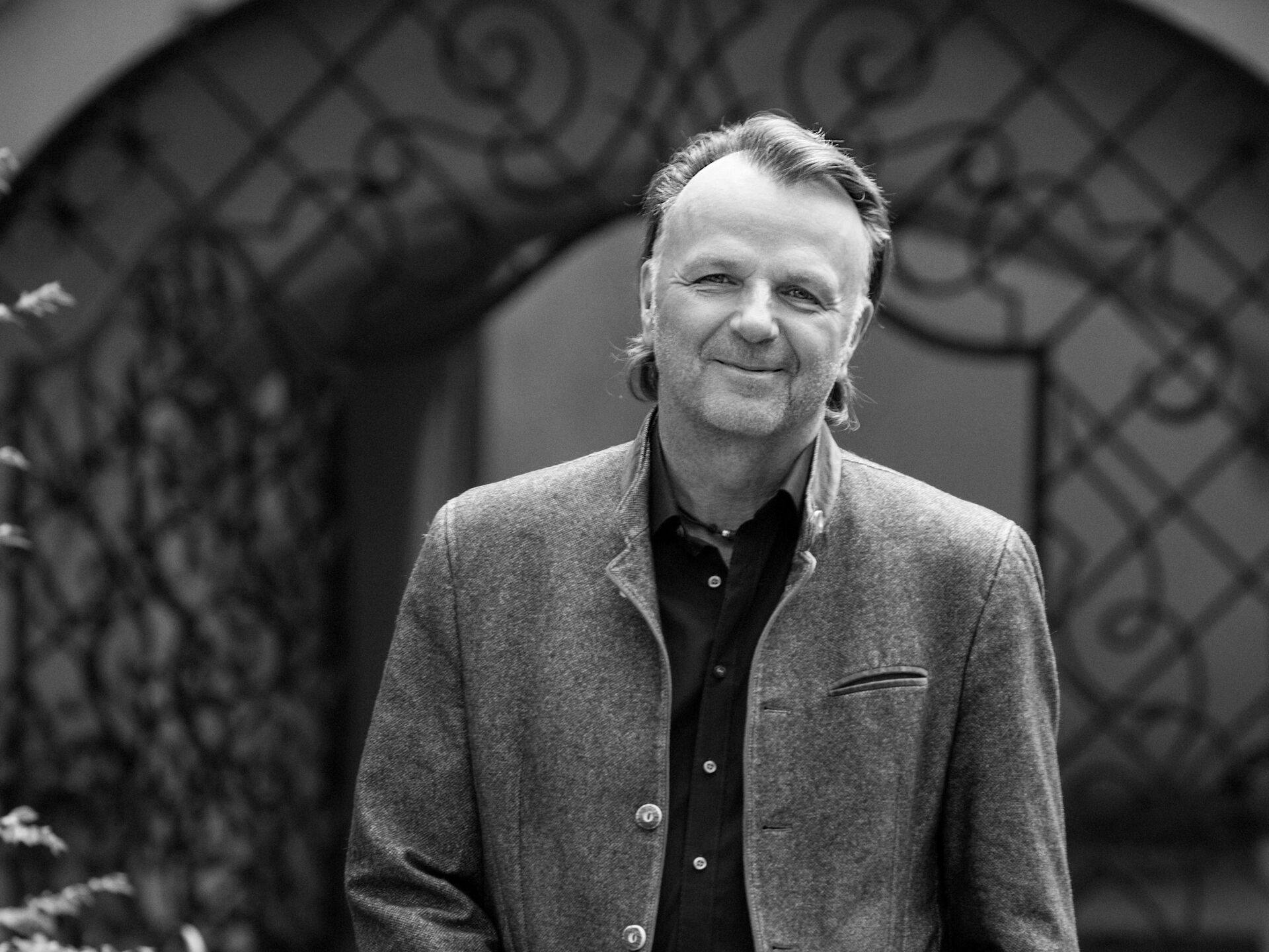 Schwarz-weiß-Foto von Jörg Brunecker in einer Freizeitjacke, der mit den Händen in den Taschen neben Gartenpflanzen und einem geschwungenen Eisengittertor steht