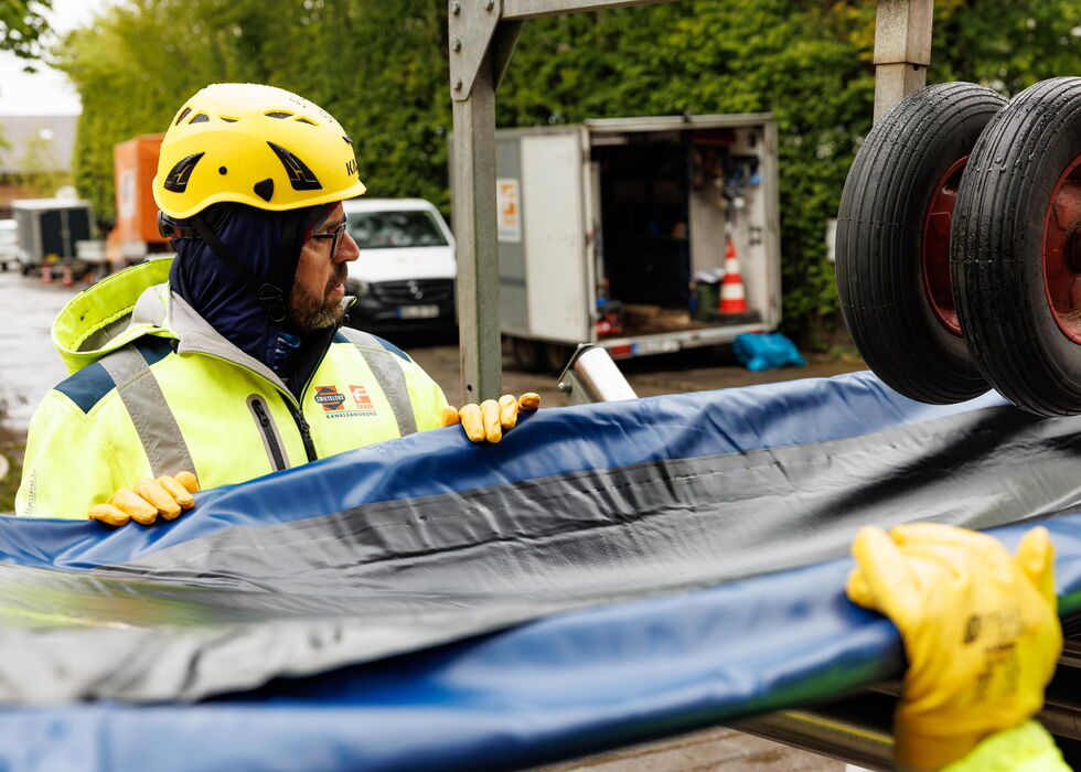 Arbeiter mit gelbem Helm und Warnjacke bewegt eine große Plane neben Lkw-Rädern an einer Straßenbaustelle.