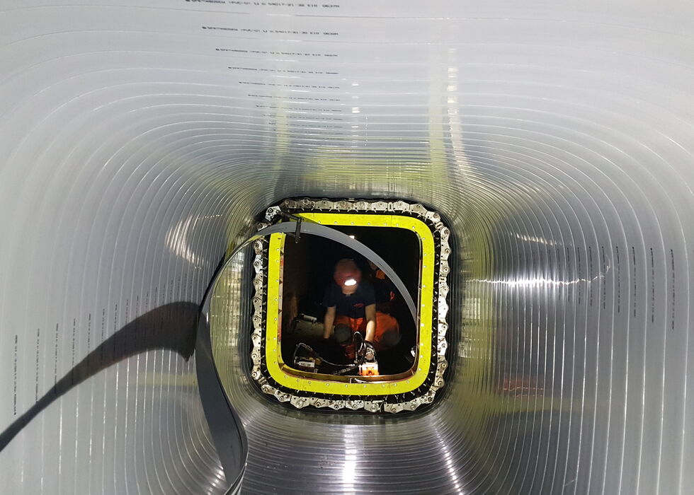 Ein Arbeiter in einer großen Metallröhre oder einem Tunnel, gesehen von innen durch die geriffelte Passage.