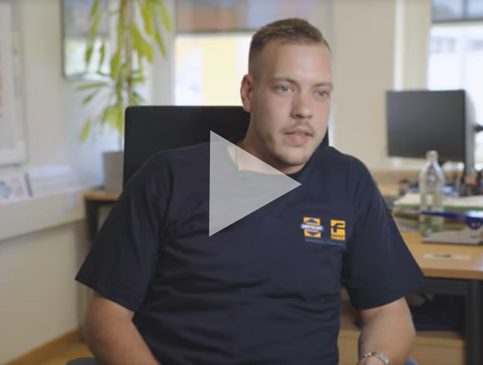 Ein Mann trägt ein dunkelblaues Hemd und sitzt in einem Büro mit Computern und Pflanzen im Hintergrund