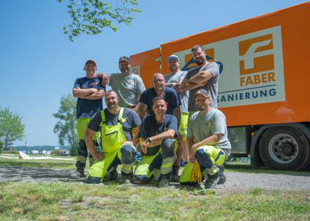 Sieben Mitarbeiter posieren im Freien neben einem orangefarbenen Transporter Swietelsky-Faber GmbH an einem sonnigen Tag.