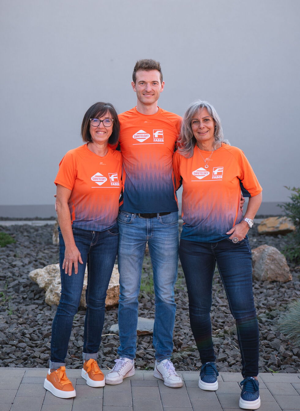 Drei Personen stehen zusammen im Freien, tragen passende orange-blaue Shirts und Jeans