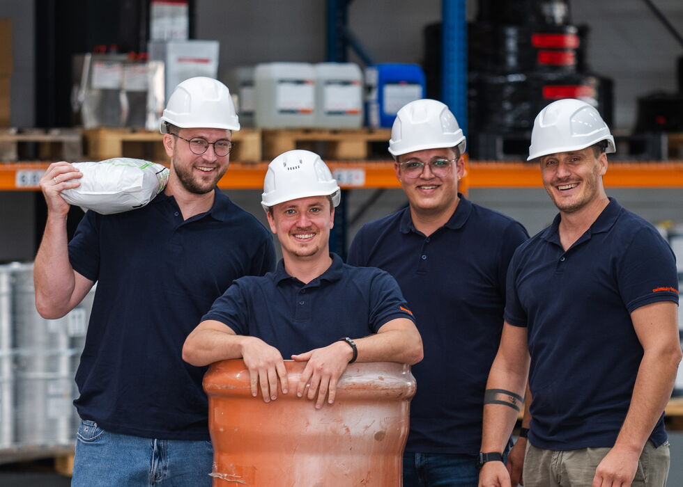 Vier Arbeiter mit Schutzhelmen stehen in einem Lager um ein großes orangefarbenes Industriefass.