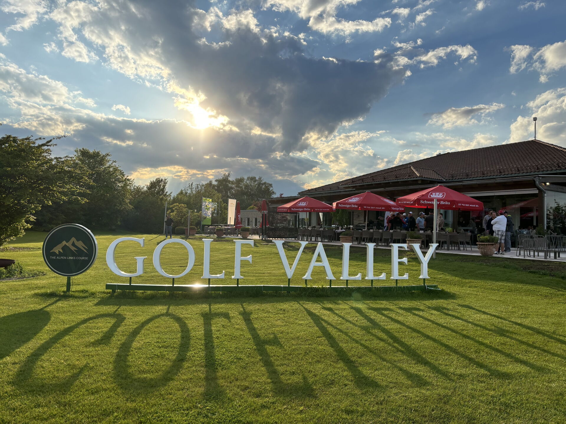 Großes Golf-Valley-Schild auf einer grünen Wiese vor Clubhaus, roten Sonnenschirmen und dramatischem Abendhimmel.
