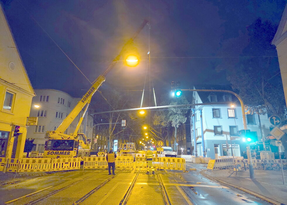 Ein Kran und Absperrungen blockieren nachts einen Straßenbahn- oder Bahnübergang, mit Straßenlaternen und einigen Personen.