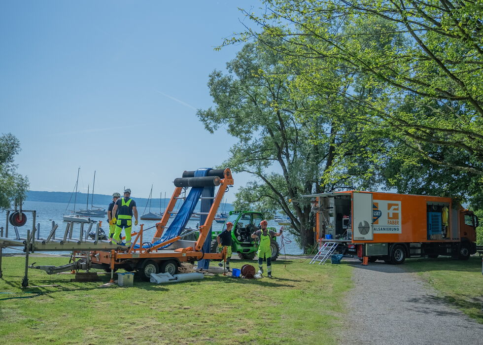 Mitarbeiter schneiden Äste mit einer Hubarbeitsbühne, während ein Versorgungs-Lkw und Werkzeuge auf einer Wiese stehen.