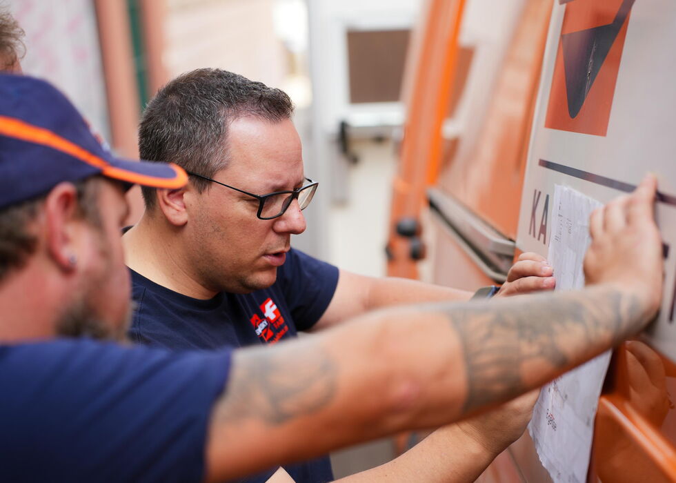 Zwei Arbeiter bringen sorgfältig Foliengrafiken an der Seite eines orangefarbenen Transporters an.