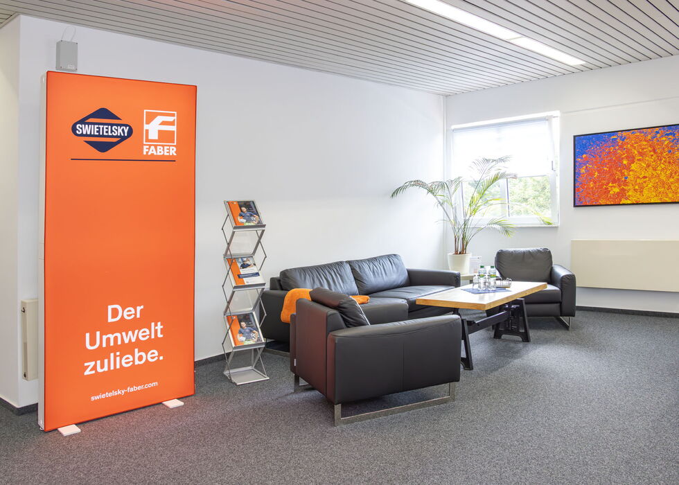 Heller Büro-Loungebereich mit schwarzen Sofas, orangefarbenem Banner, Prospektständer, Tisch und Wandbild.
