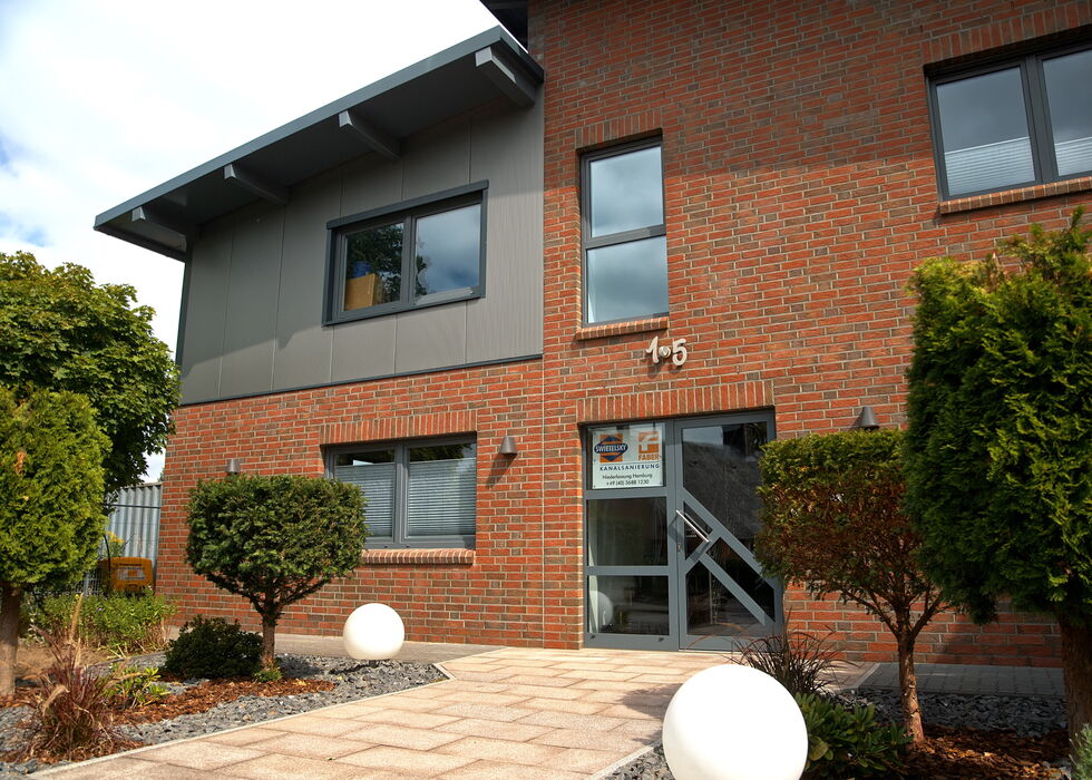 Exterior of a modern brick office building with windows, shrubs, and a paved entrance walkway.