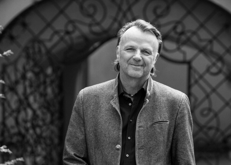 Schwarz-weiß-Foto von Jörg Brunecker in einer Freizeitjacke, der mit den Händen in den Taschen neben Gartenpflanzen und einem geschwungenen Eisengittertor steht