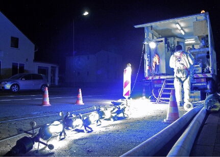 Ein Arbeiter bedient Geräte bei Nacht in einer hell erleuchteten Baustelle mit Pylonen und Maschinen.