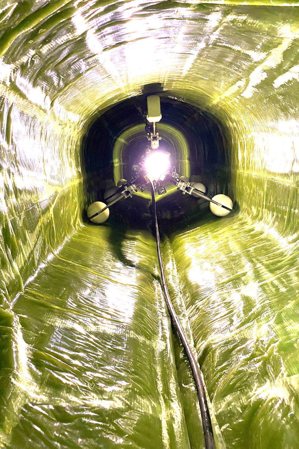 Blick in einen glänzenden gelb-grünen Industrietunnel mit einem Kabel, das zu einem hellen Licht führt.