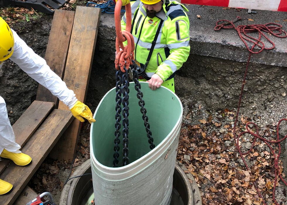 Bauarbeiter in Schutzausrüstung senkt Ketten und eine Pumpe in einen Versorgungsschacht an einer Straßenbaustelle.