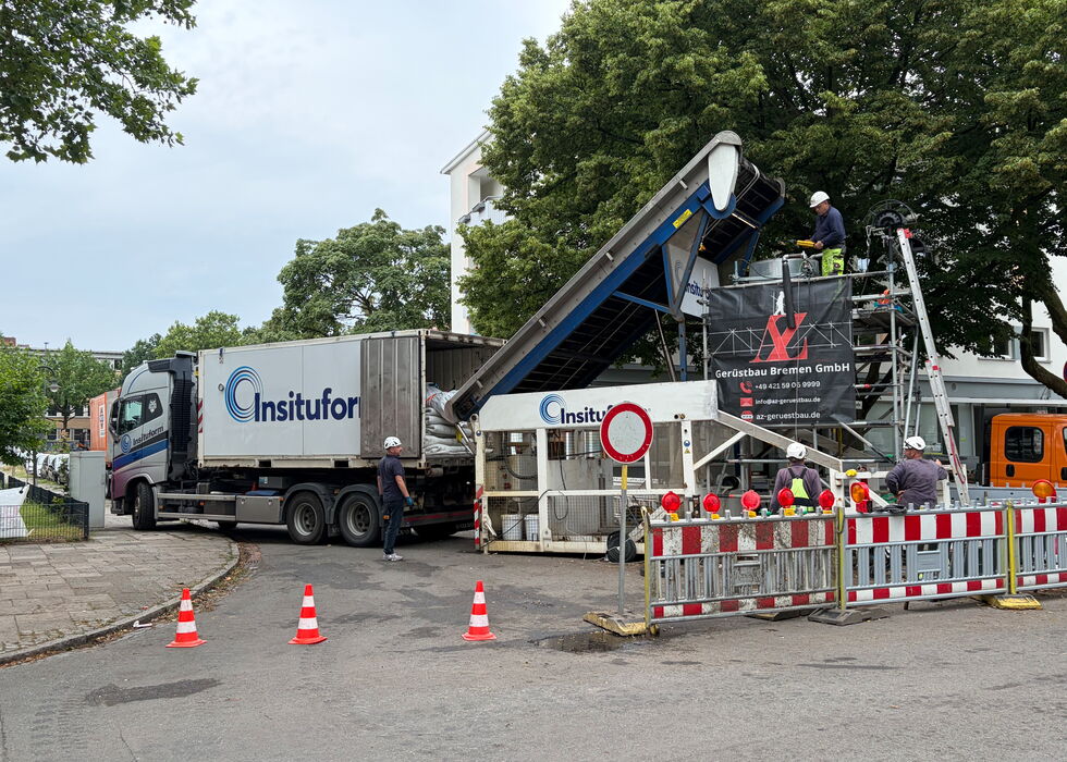 Lkw lädt Synthesefaserliners auf einer Baustelle mit Arbeitern, Absperrungen und Pylonen.