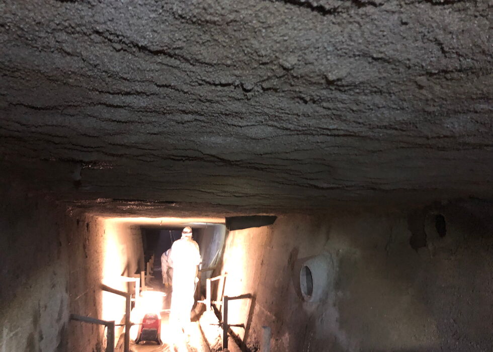 Person in Schutzanzug steht in einem schlecht beleuchteten, engen Tunnel mit Betonwänden und -decke