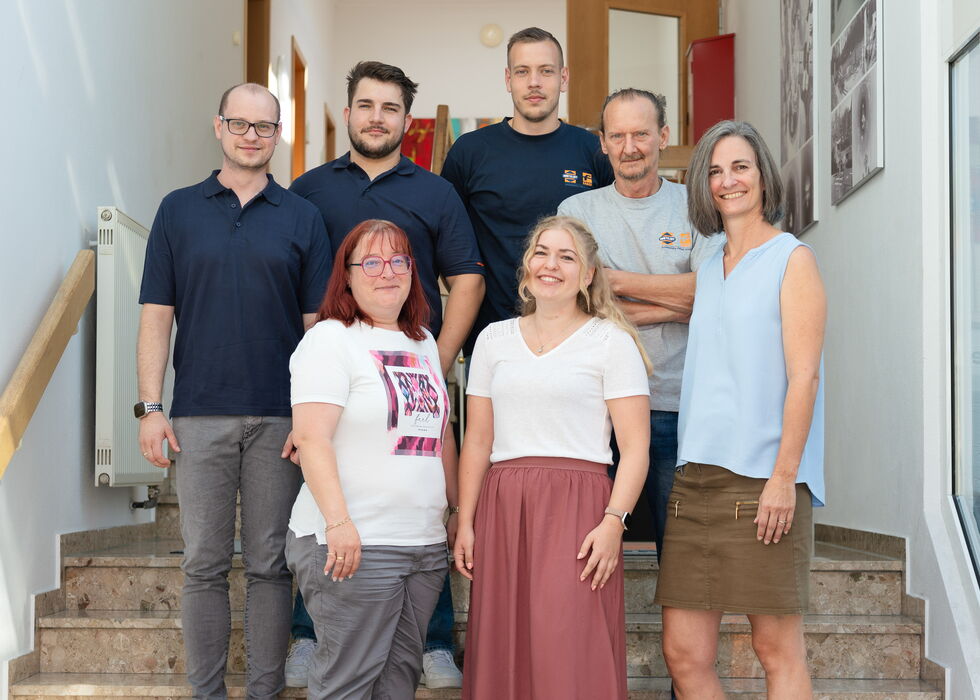Sieben Personen posieren gemeinsam auf einer Innentreppe und lächeln in die Kamera.