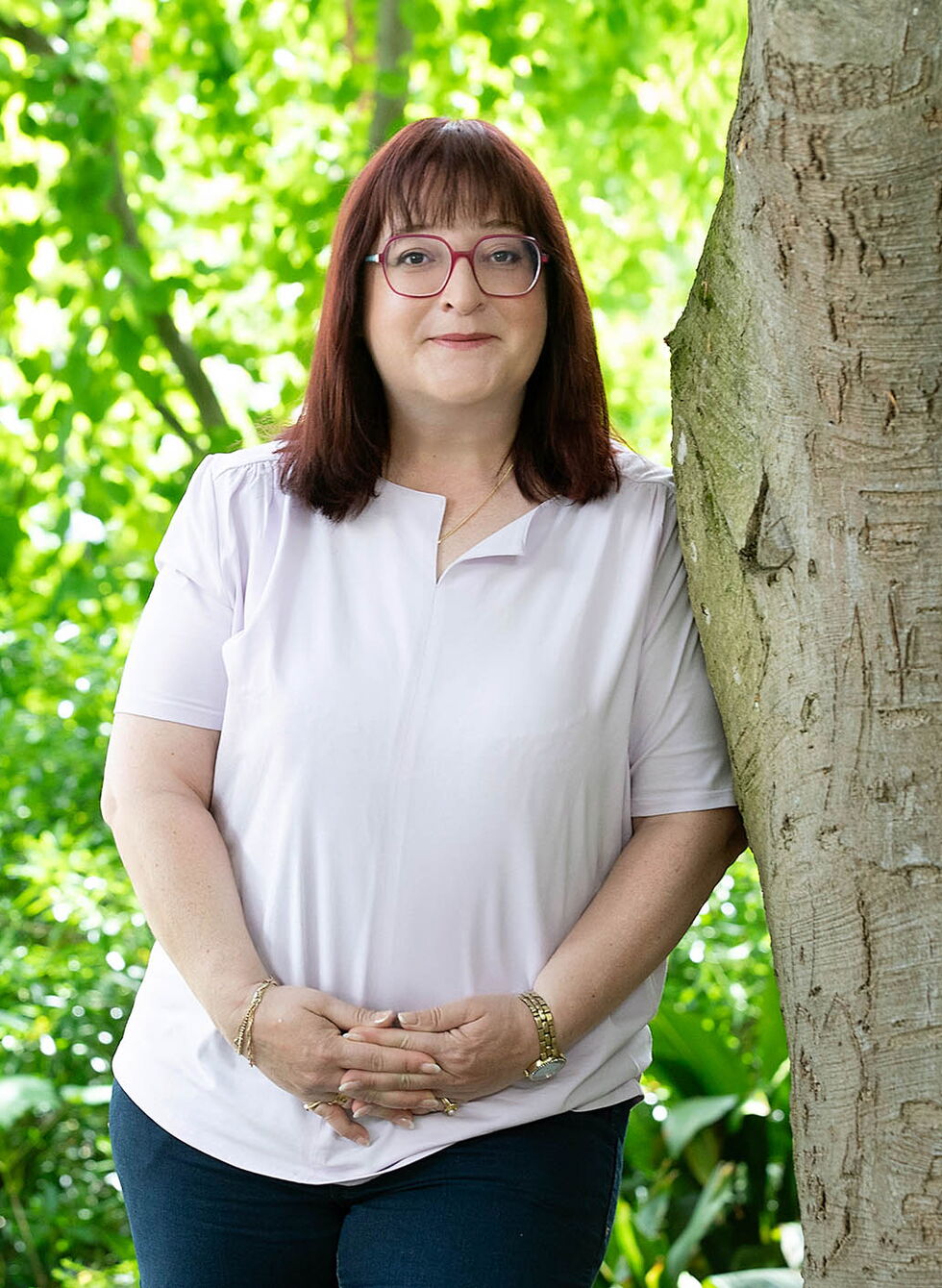 Eine Frau mit Brille steht neben einem Baum in einem grünen Park.