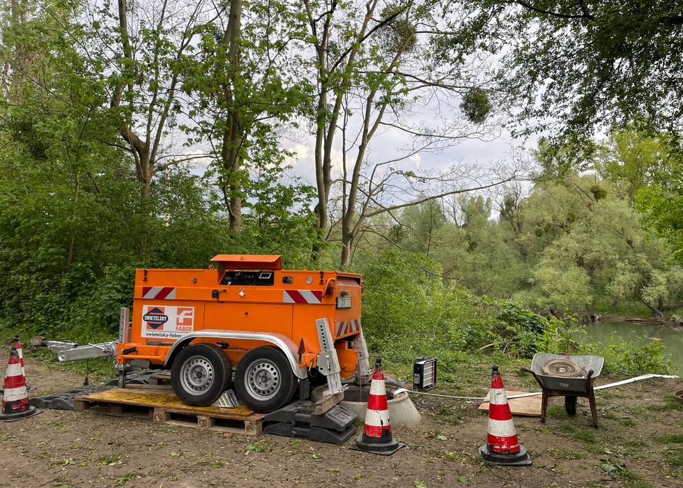 Orangefarbener Arbeitsanhänger auf einer Plattform am Flussufer, umgeben von Bäumen und Leitkegeln.