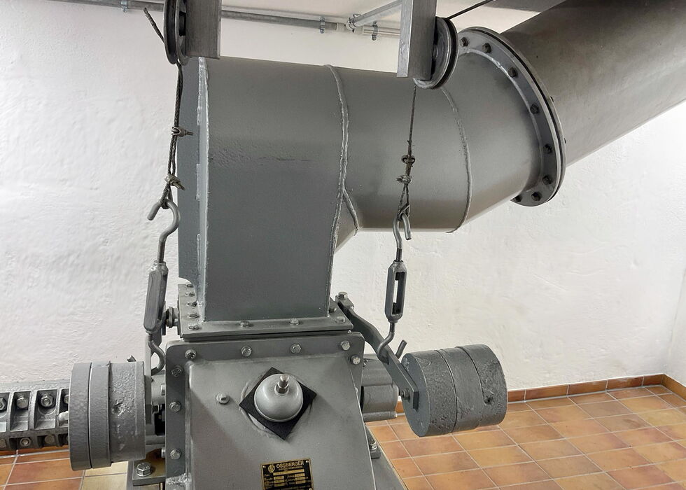 Nahaufnahme einer grauen Industriemaschine, die in einem gefliesten Raum mit großen Lüftungskanälen verbunden ist.