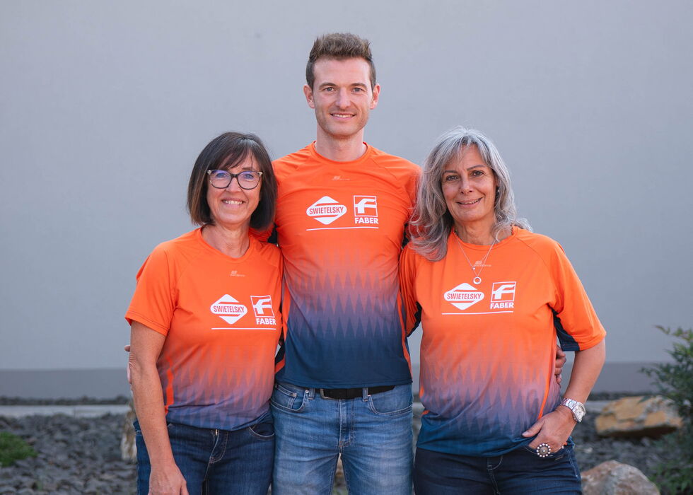 Drei lächelnde Personen stehen draußen zusammen und tragen passende orangefarbene Teamshirts und Jeans.