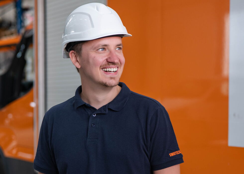 Lächelnder Mann mit Schutzhelm und dunkelblauem Poloshirt steht neben einem orangefarbenen Lkw in einem Lager.