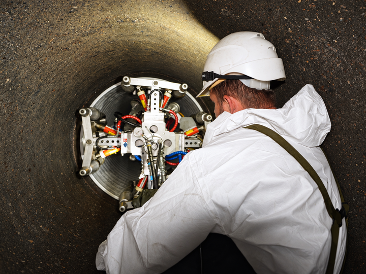 A worker wearing a protective suit and helmet is inspecting the winding pipe system inside a large sewer pipe.