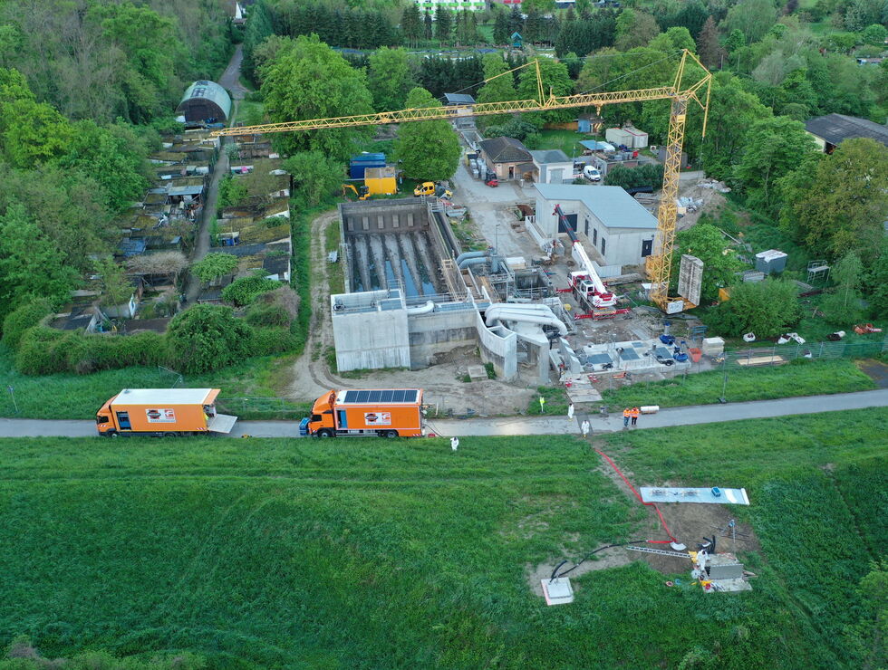 Luftaufnahme einer Baustelle mit Kran, im Bau befindlichen Gebäuden und Lastwagen neben einer Straße, umgeben von Feldern und Bäumen.