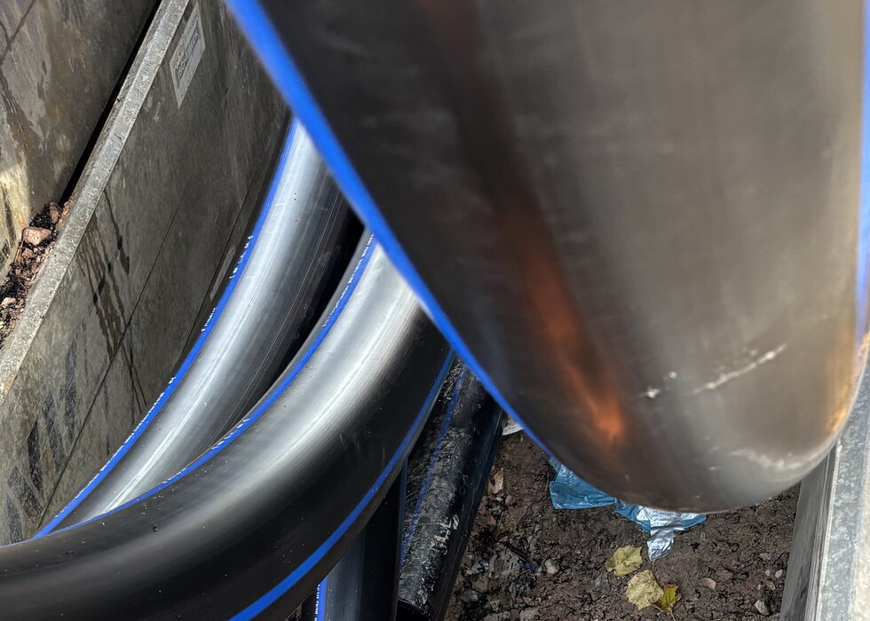 Nahaufnahme schwarzer Kabel mit blauen Streifen, verlegt in einem engen Graben neben einem großen Rohr.