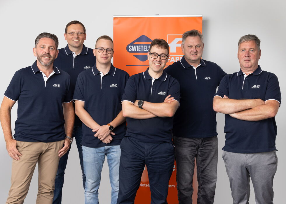 Sechs Teammitglieder stehen vor einem orangefarbenen Banner für ein Gruppenfoto.