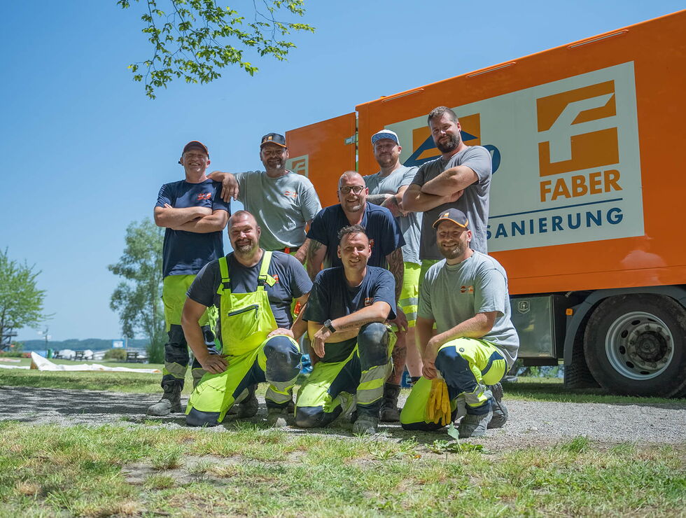 Gruppe von Mitarbeitenden posiert vor einem orangefarbenen Lkw im Freien bei Sonnenschein.