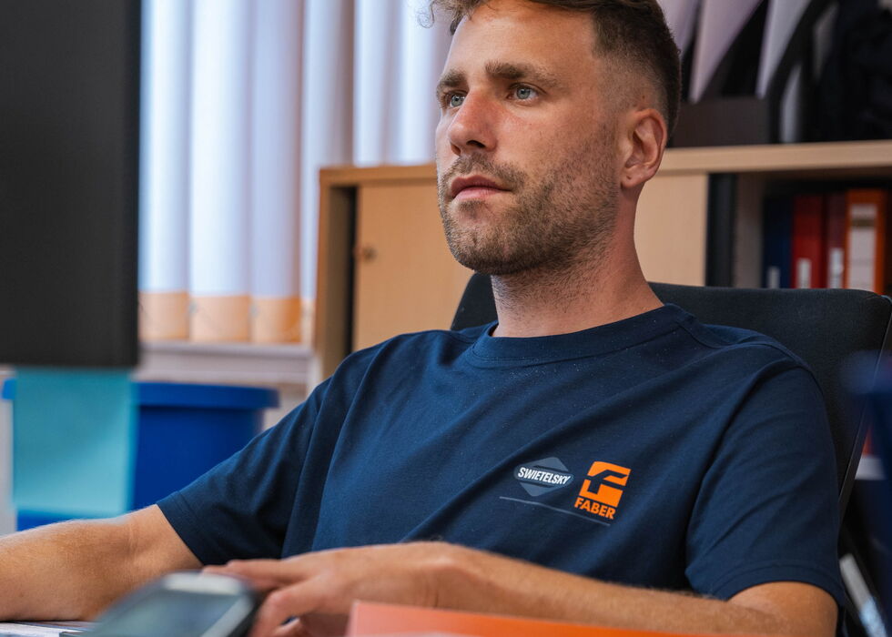 Ein Mann im dunkelblauen T-Shirt sitzt an einem Schreibtisch im Büro, im Hintergrund Bücher und Unterlagen.