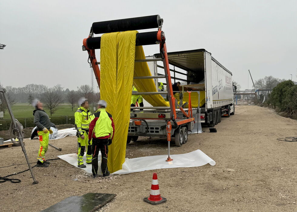 Arbeiter in Warnkleidung entladen eine große gelbe Geberolle neben einer Landstraße aus einem Lkw.